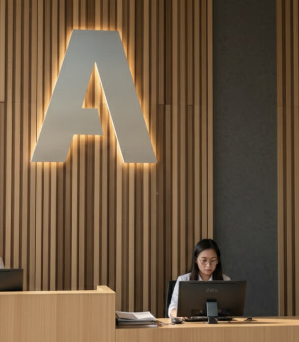 An office reception desk with a woman behind the desk working on a computer. A large Authentia logo can be seen on the wall behind her.